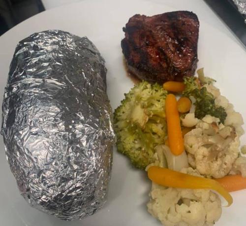 A plate with a foil-wrapped baked potato, grilled steak, and a mix of broccoli, cauliflower, and carrots.