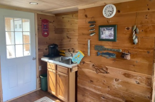 A cozy wooden kitchen corner with a sink, clock, and fishing-themed decor, featuring a door leading outside.