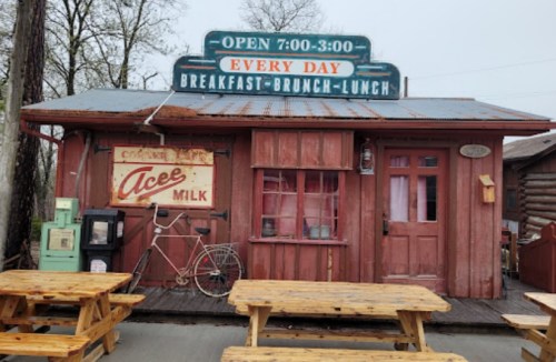 A rustic diner with a red wooden exterior, picnic tables outside, and a sign indicating breakfast, brunch, and lunch hours.