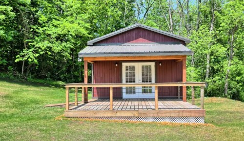 A small red cabin with a wooden deck, surrounded by lush green trees and grass.