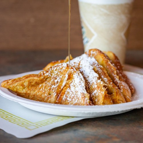 A plate of French toast slices drizzled with syrup and dusted with powdered sugar, with a drink in the background.