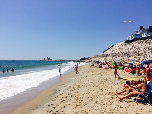 A sunny beach scene with people swimming, sunbathing, and a rocky shoreline under a clear blue sky.