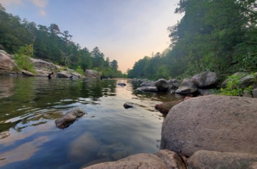 A serene river scene surrounded by trees and rocks, reflecting the sky at sunset.