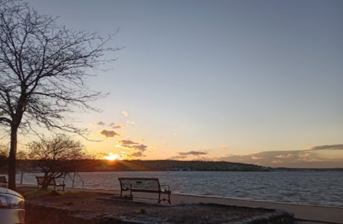 A serene lakeside view at sunset, featuring a bench, trees, and calm water reflecting the colorful sky.