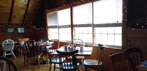 Cozy restaurant interior with wooden decor, tables, chairs, and warm lighting from string lights by large windows.