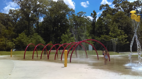 A child plays in a splash pad with red arches and water sprays, surrounded by trees and a blue sky.