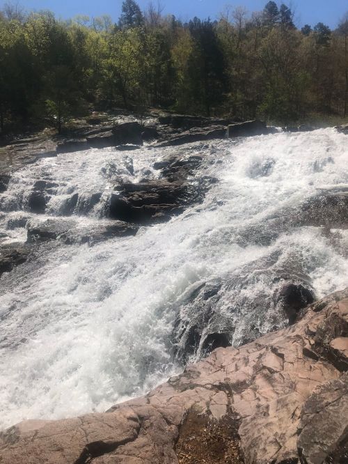 A rushing waterfall cascades over rocky terrain, surrounded by lush green trees under a clear blue sky.