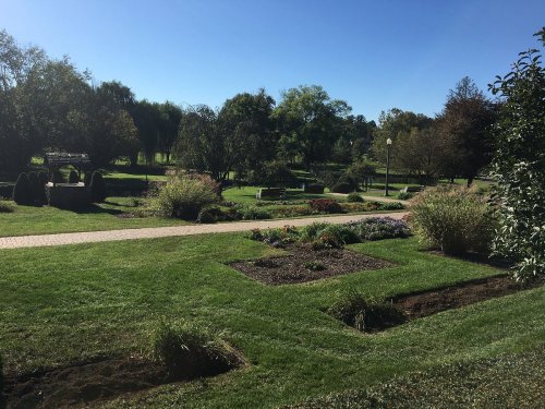 A sunny park scene with green lawns, flower beds, and walking paths surrounded by trees and benches.
