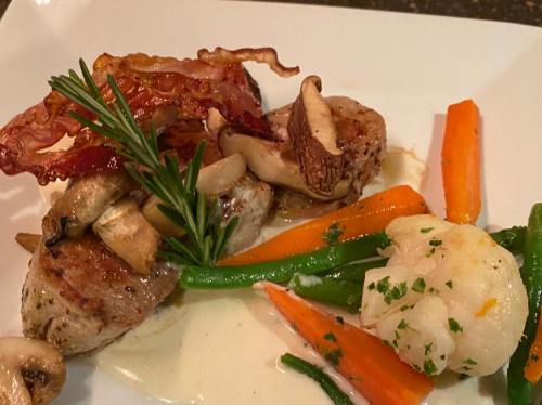 A gourmet plate featuring seared meat, sautéed mushrooms, crispy bacon, and colorful vegetables on a white dish.
