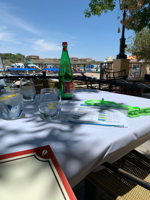 A table set for dining outdoors, featuring glasses of water, a bottle of soda, and a menu, with a view of the water.