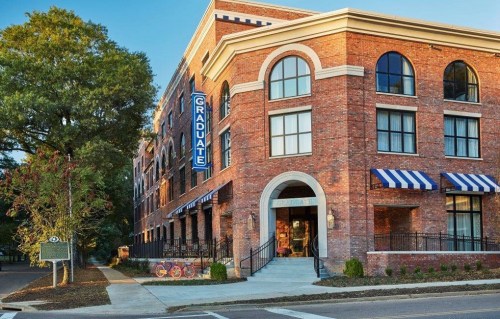A brick hotel building with large windows and a "Graduate" sign, featuring striped awnings and surrounding greenery.