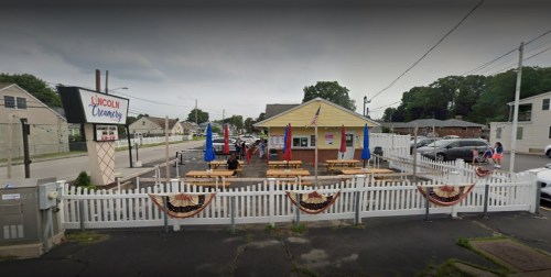 A small restaurant with outdoor seating, flags, and a white picket fence, set in a suburban area.