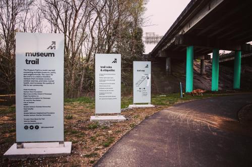 Signage for a museum trail under a bridge, with information about trail rules and directions. A dog is visible nearby.
