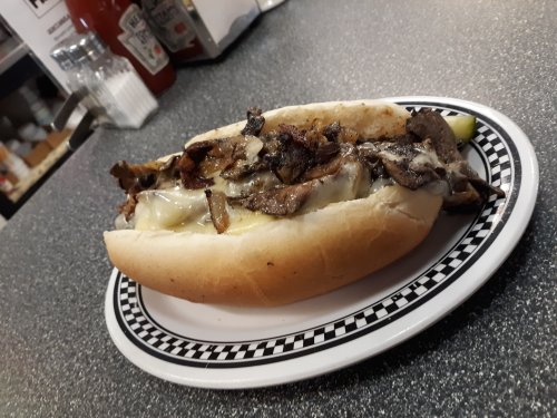 A loaded sandwich with grilled beef, melted cheese, and toppings, served in a soft bun on a checkered plate.
