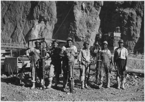 Black and white photo of six miners posing with tools in a rocky landscape, showcasing their work environment.