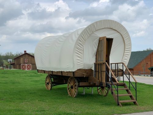 A covered wagon on wheels, parked on green grass with a barn in the background under a cloudy sky.