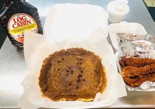 A large pancake topped with chocolate chips, served with bacon and a bottle of syrup on the side.