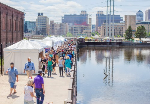 A bustling outdoor event along a waterfront, with crowds walking past white tents and a heron in the water.