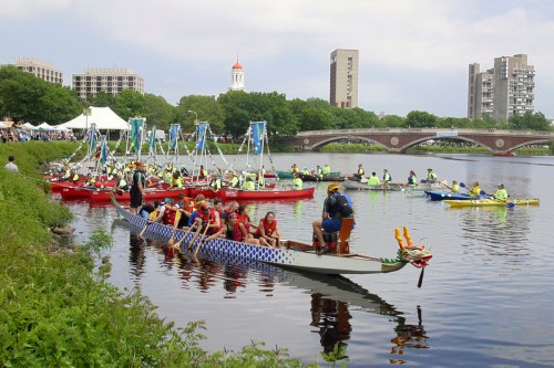 A vibrant scene of dragon boat teams preparing on a river, with buildings and greenery in the background.