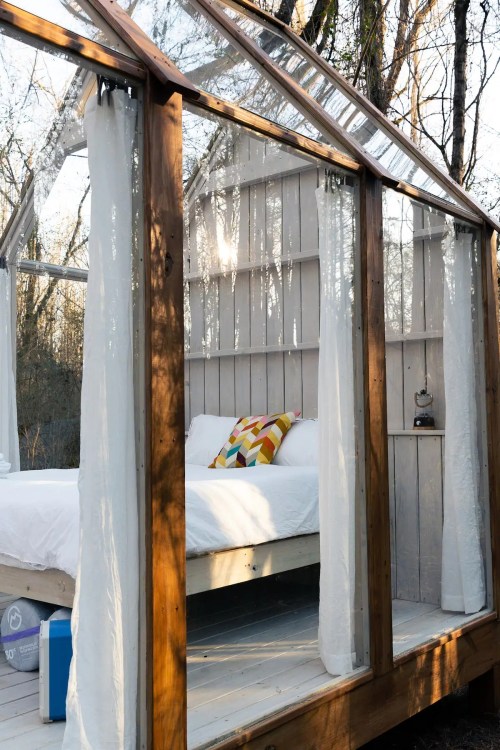 A cozy, transparent cabin with a bed and colorful pillows, surrounded by trees in a serene outdoor setting.