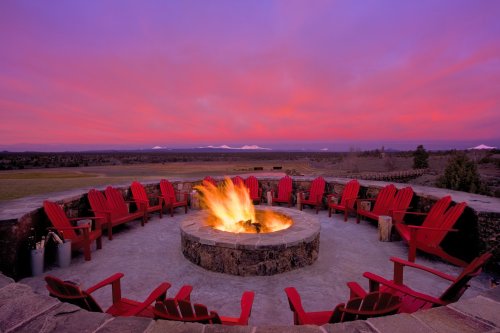 A circular stone fire pit surrounded by red chairs, set against a vibrant sunset sky and distant mountains.