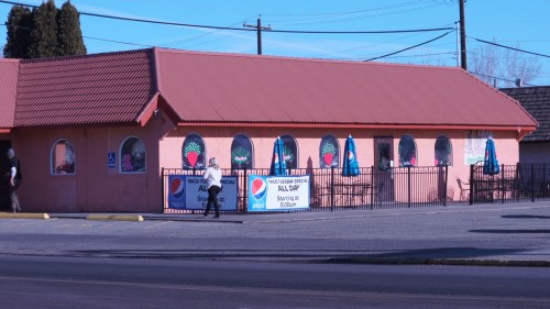 A pink building with a red roof, featuring large windows and outdoor seating, surrounded by banners and a parking lot.