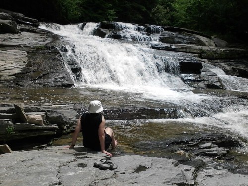 A person in a white hat sits on rocks by a waterfall, surrounded by lush greenery and flowing water.