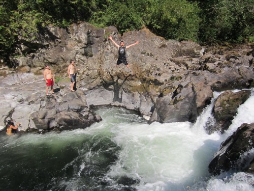 A person jumps off a rock into a river while others stand nearby, surrounded by lush greenery and rocky terrain.
