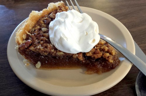 A slice of pecan pie topped with whipped cream on a plate, with a fork and spoon beside it.