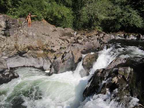 A person stands on a rocky riverbank, pointing towards a rushing river surrounded by lush greenery.