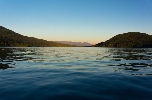 Serene lake view at sunset, surrounded by mountains and a clear sky, reflecting calm water.