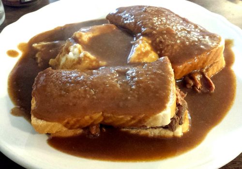 A plate of sliced bread topped with gravy, served alongside mashed potatoes and meat.