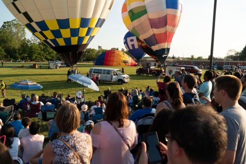 A crowd watches as colorful hot air balloons prepare for launch at a festival on a sunny day.
