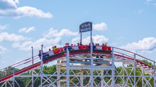 A group of people rides a wooden roller coaster under a blue sky with fluffy clouds.