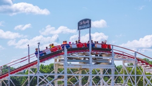A wooden roller coaster with riders at the top, set against a blue sky with fluffy clouds.