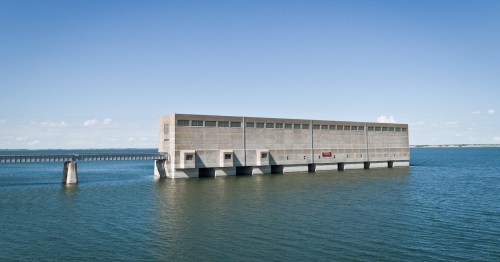 A large, modern concrete building stands on stilts over calm water, with a clear blue sky in the background.