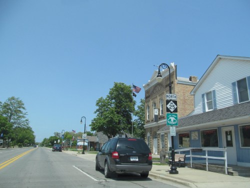 A small town street with a black minivan, street signs, and buildings under a clear blue sky.