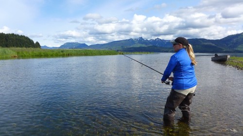 A person fishing in a river surrounded by mountains and greenery, wearing a blue jacket and waders.
