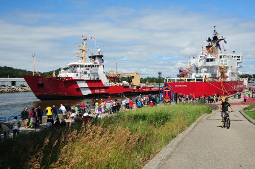 A red ship docked with people gathered along the waterfront, enjoying a sunny day by the water.