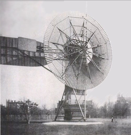 A large, vintage wind turbine with a circular blade structure, set in a grassy area with trees in the background.