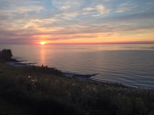 A serene sunset over calm waters, with silhouettes of people on the shore and colorful clouds in the sky.