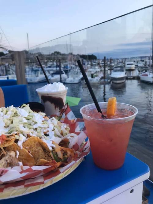 A plate of nachos and a cocktail sit on a table with a marina view in the background during sunset.