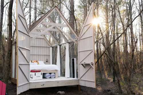 A minimalist open-air cabin with a bed, surrounded by trees, and sunlight streaming through the branches.