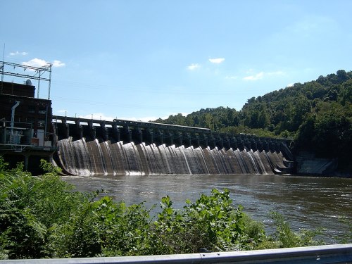 A dam with water cascading over its structure, surrounded by greenery and a clear blue sky.