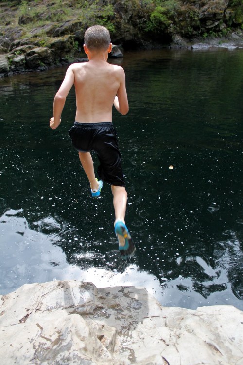 A boy jumps off a rock into a dark pool of water, creating splashes around him.