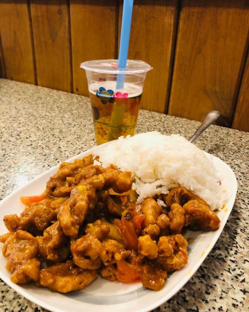 A plate of orange chicken with rice and a cup of iced tea on a speckled table.