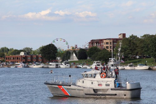 A U.S. Coast Guard boat navigates a river with a ferris wheel and buildings in the background on a sunny day.