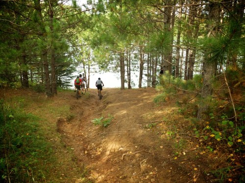 Two cyclists ride down a dirt path through a forest, approaching a calm lake surrounded by trees.