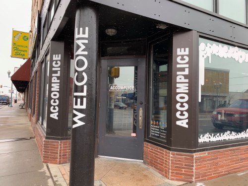 Exterior of a storefront with "WELCOME" and "ACCOMPLICE" signs on the windows and door, featuring a brick facade.