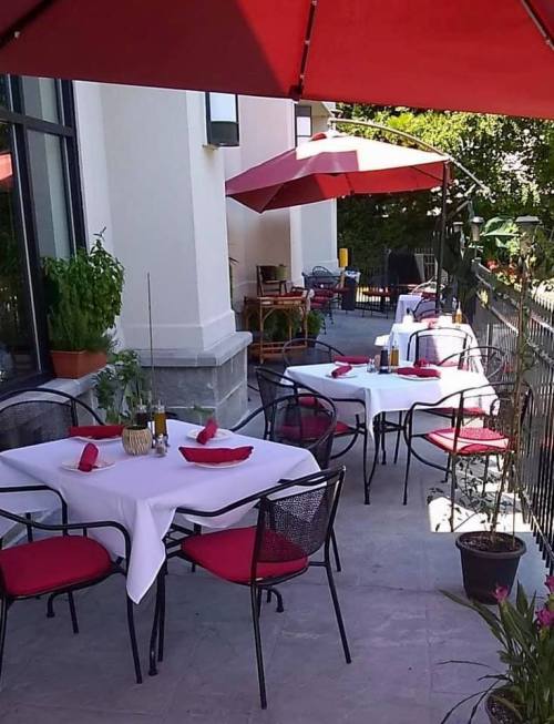 Outdoor dining area with tables set for guests, featuring red umbrellas and potted plants.